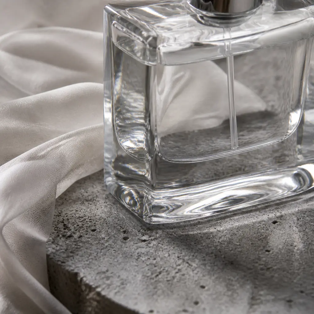 Extreme close-up of a clear perfume bottle refracting a rough stone slab beside draped translucent silk